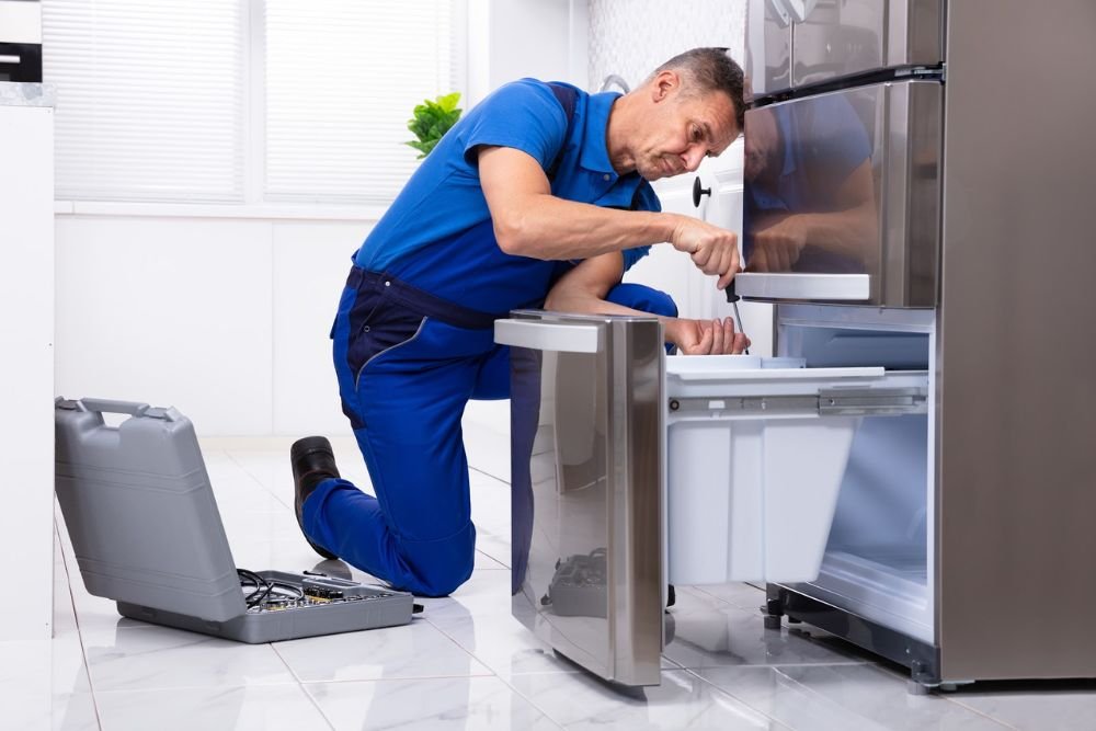 A technician in blue overalls works on a refrigerator, showcasing a kitchen appliance repair service in Reno, NV.